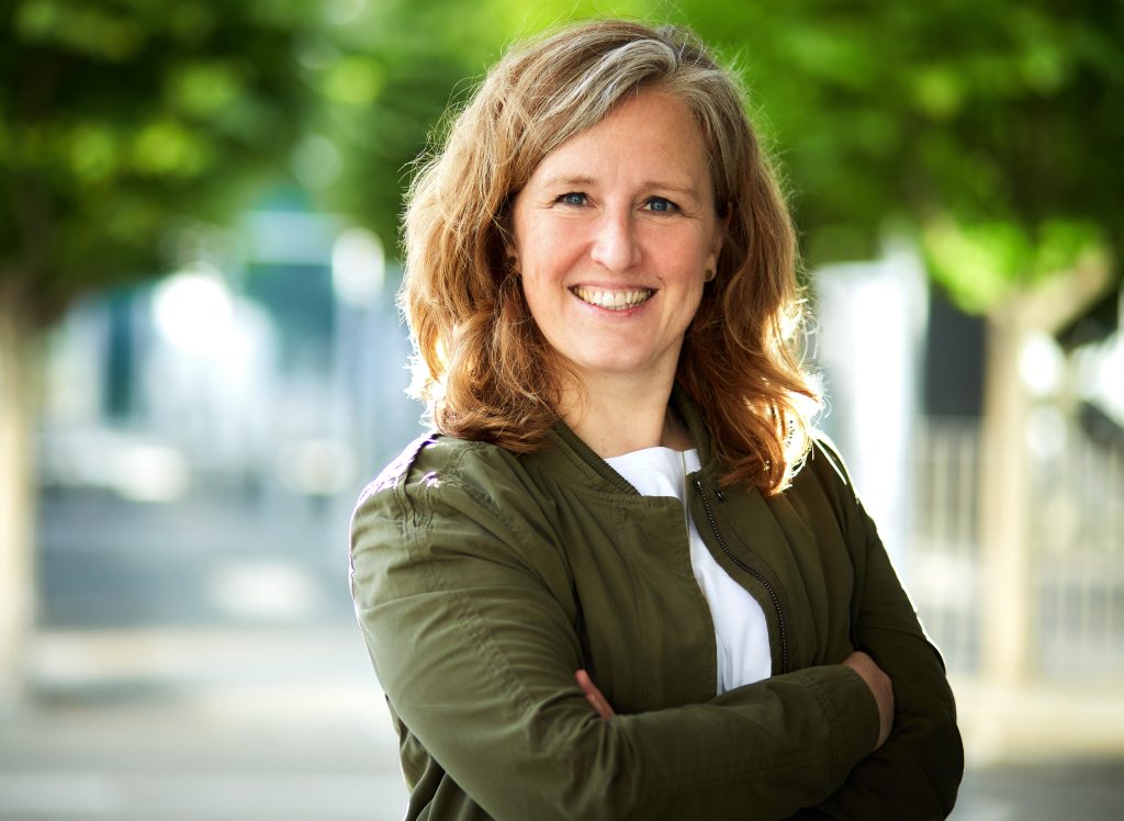 Eine lächelnde Frau mittleren Alters mit schulterlangen, hellbraunen Haaren steht im Freien vor einem unscharfen, grünen Hintergrund. Sie trägt eine olivgrüne Jacke über einem weißen Oberteil und hat die Arme locker vor der Brust verschränkt. Die Aufnahme wirkt freundlich und professionell, mit natürlichem Licht und einer entspannten Atmosphäre.