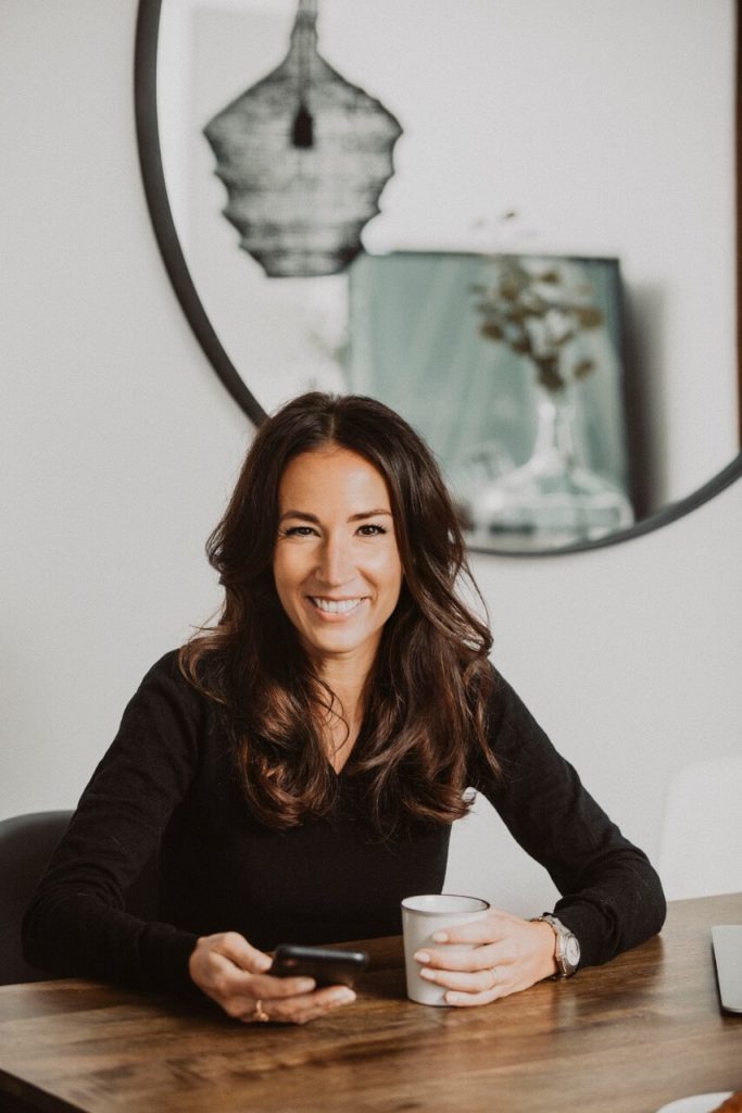 Eine lächelnde Frau mit langen, dunkelbraunen Haaren sitzt an einem Holztisch und blickt direkt in die Kamera. In einer Hand hält sie ein Smartphone, in der anderen eine helle Tasse. Sie trägt ein schwarzes Oberteil und eine Armbanduhr. Im Hintergrund ist eine helle Wand mit einem runden Spiegel sowie ein dekoratives Bild zu sehen.